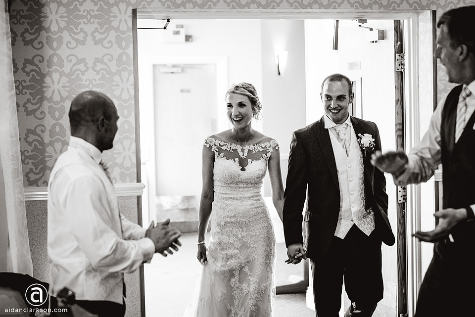 A black and white wedding photo of a bride and groom walking into a room at Kenwick Park.