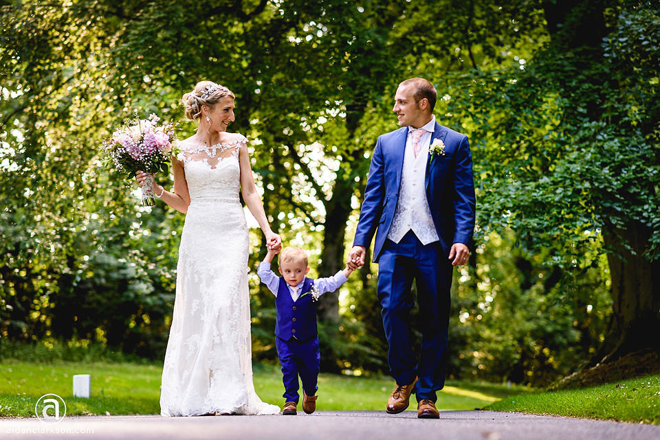 A bridal couple strolling down a path at Kenwick Park on their wedding day with their son by their side.