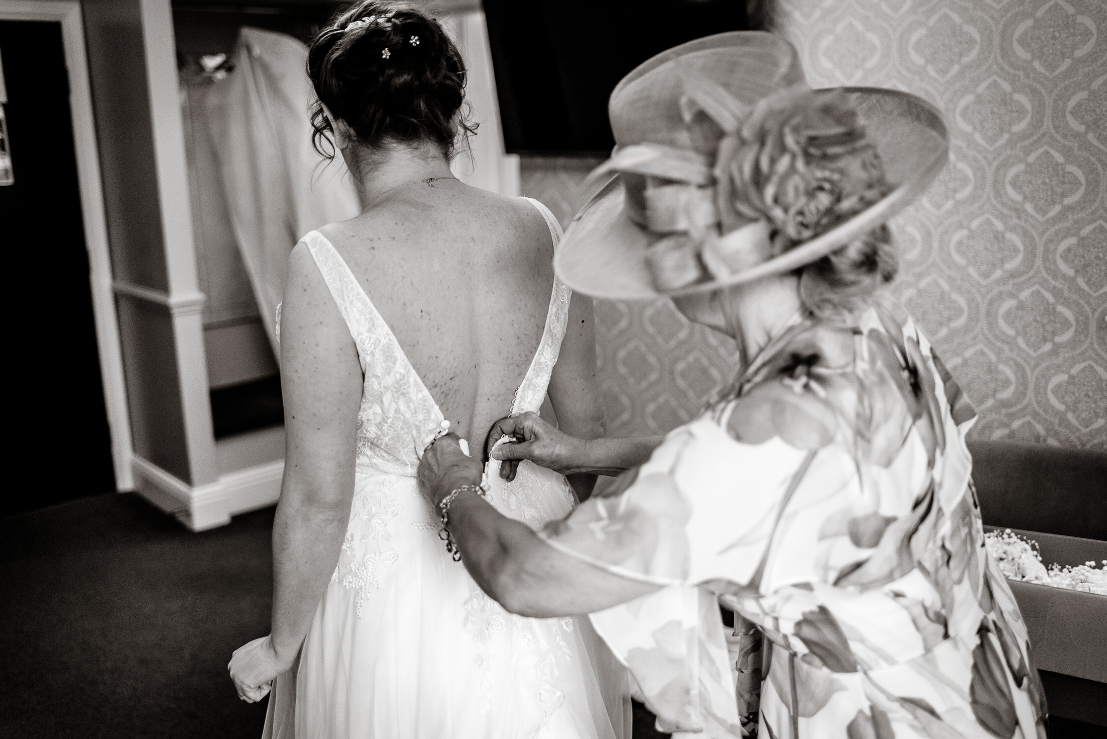 At the Brackenborough Hotel, a woman is assisting a bride in putting on her exquisite wedding dress.