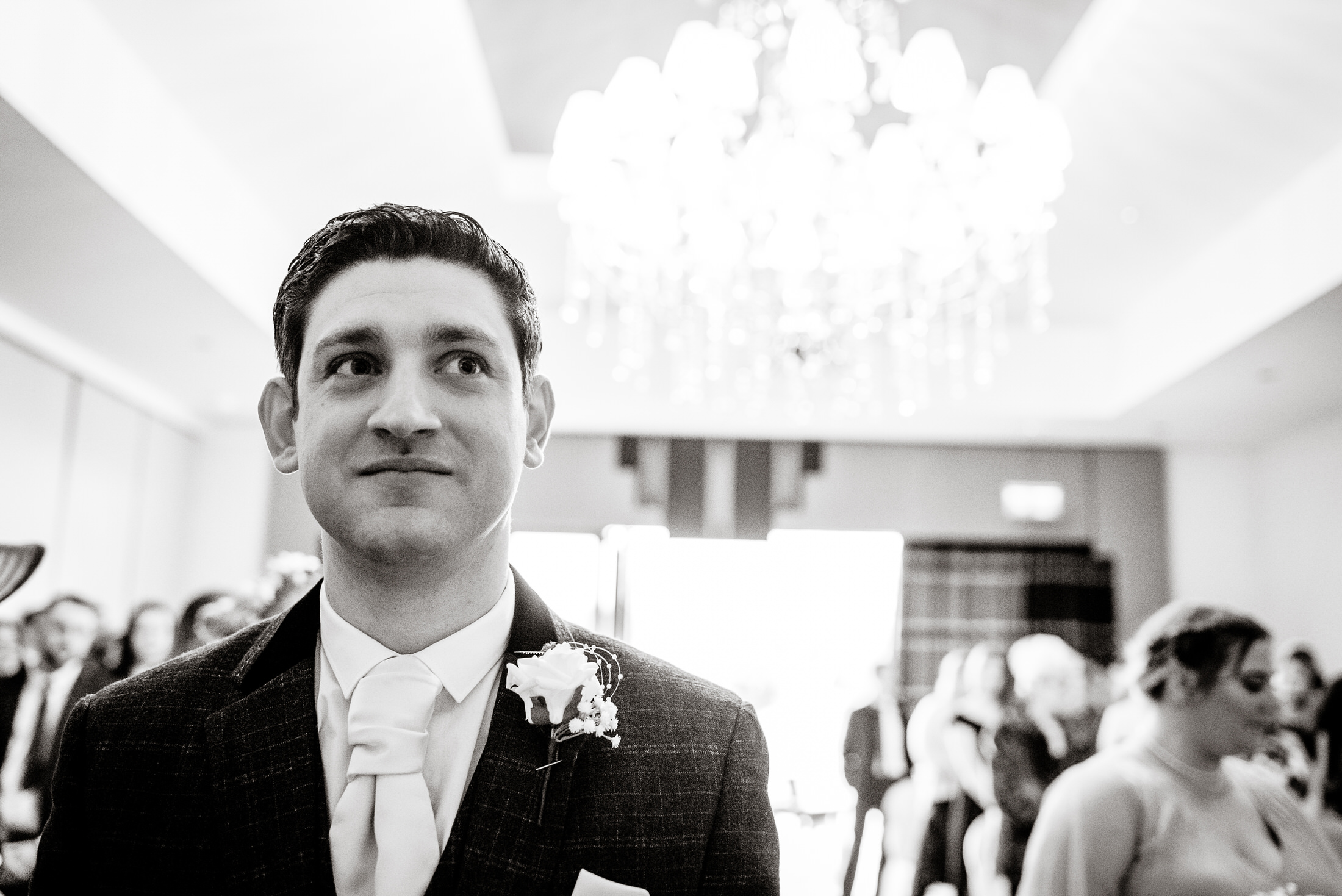 A black and white photo of a man standing in front of a chandelier at Brackenborough Hotel.