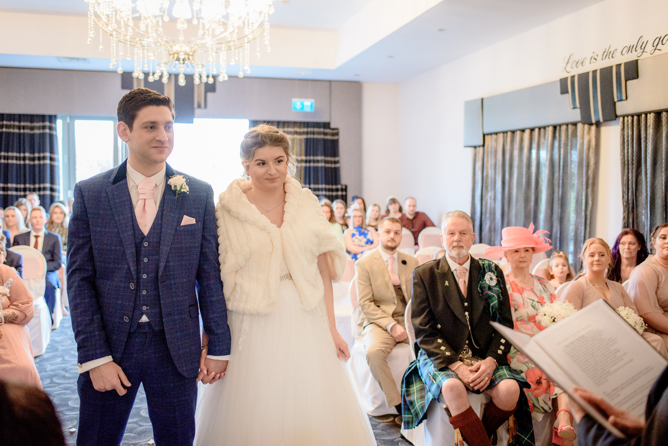 A couple, adorned in elegant attire, joyfully strolls down the aisle of the Brackenborough Hotel during their beautiful wedding ceremony.