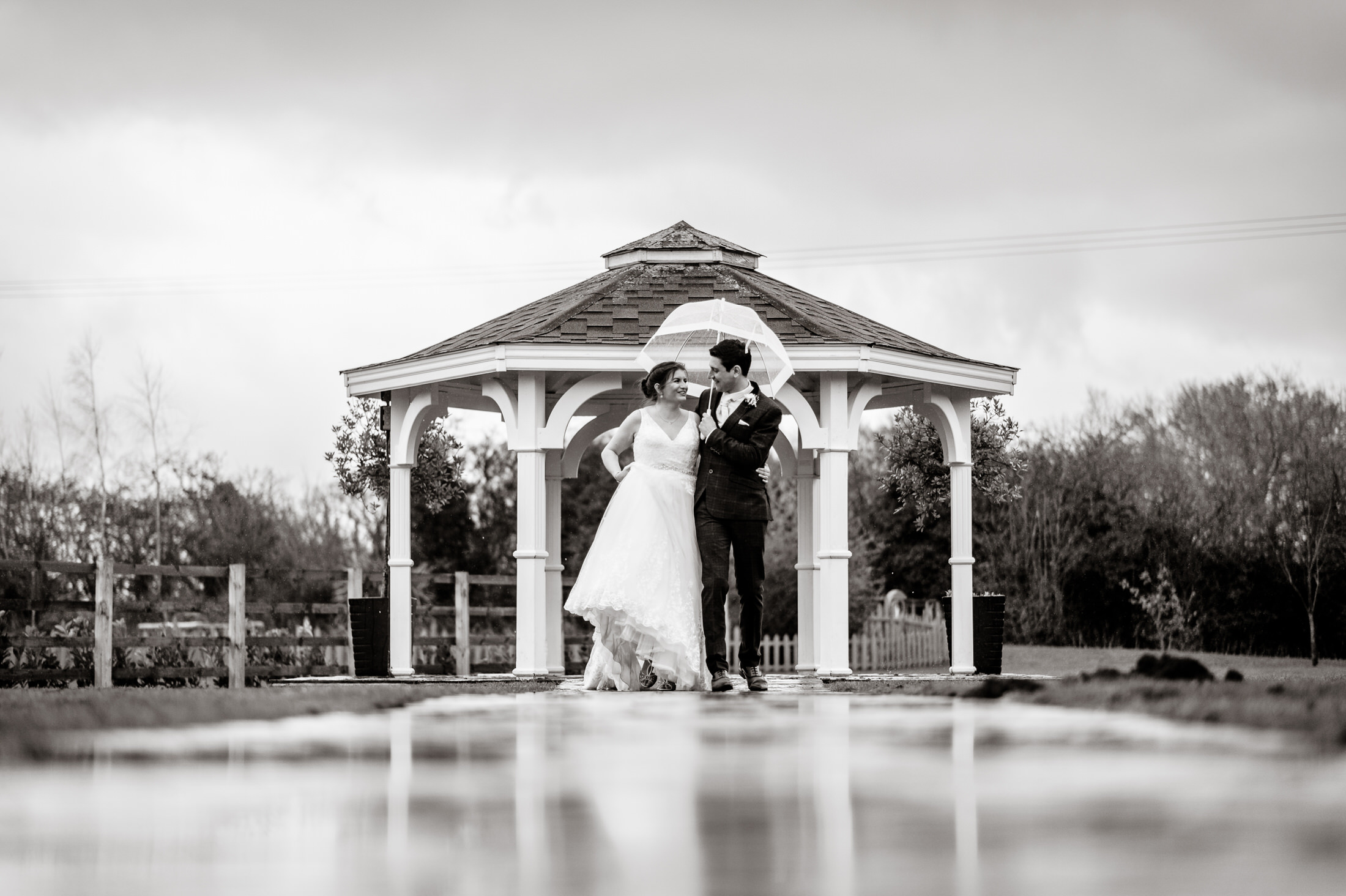 A bride and groom standing under an umbrella in front of the Brackenborough Hotel gazebo during their wedding.