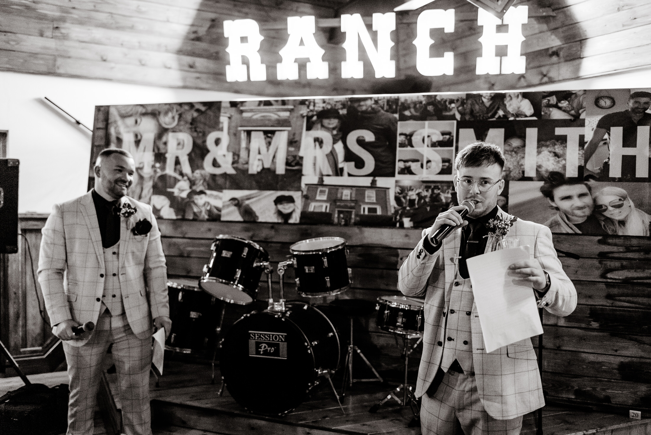 Two men in suits standing in front of a microphone at a wedding.