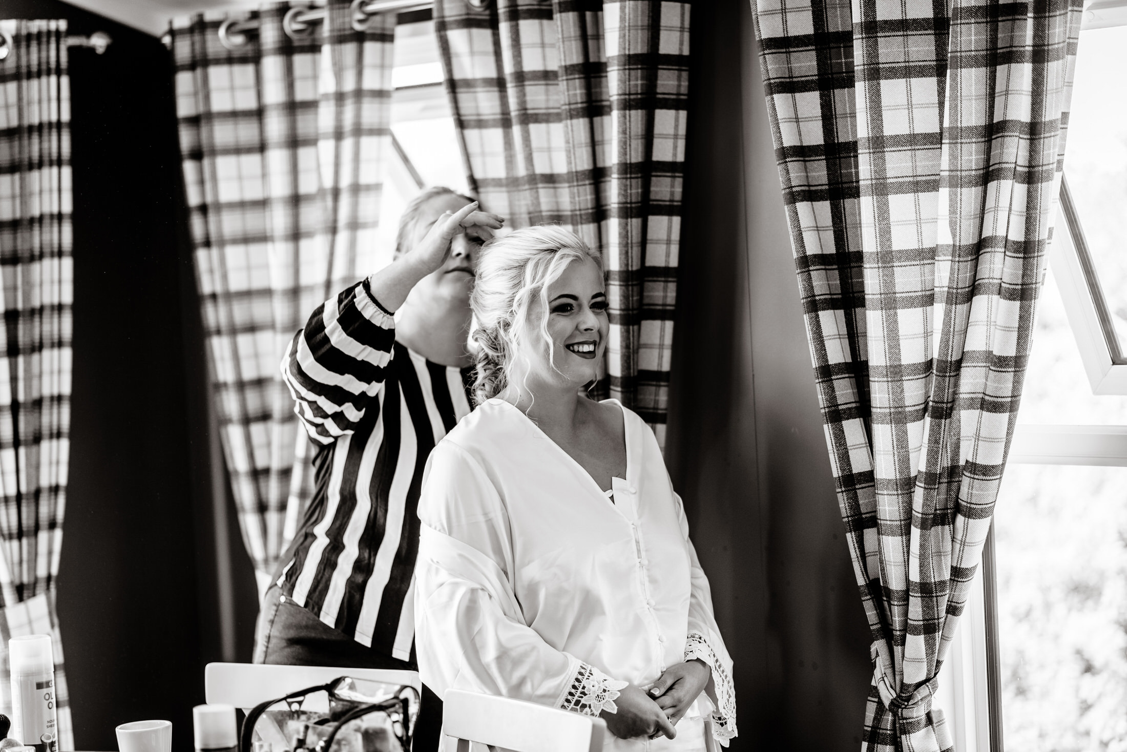 A woman in a white robe stands smiling as another woman adjusts her hair, framed by the plaid curtains reminiscent of those at Brackenborough Hotel.