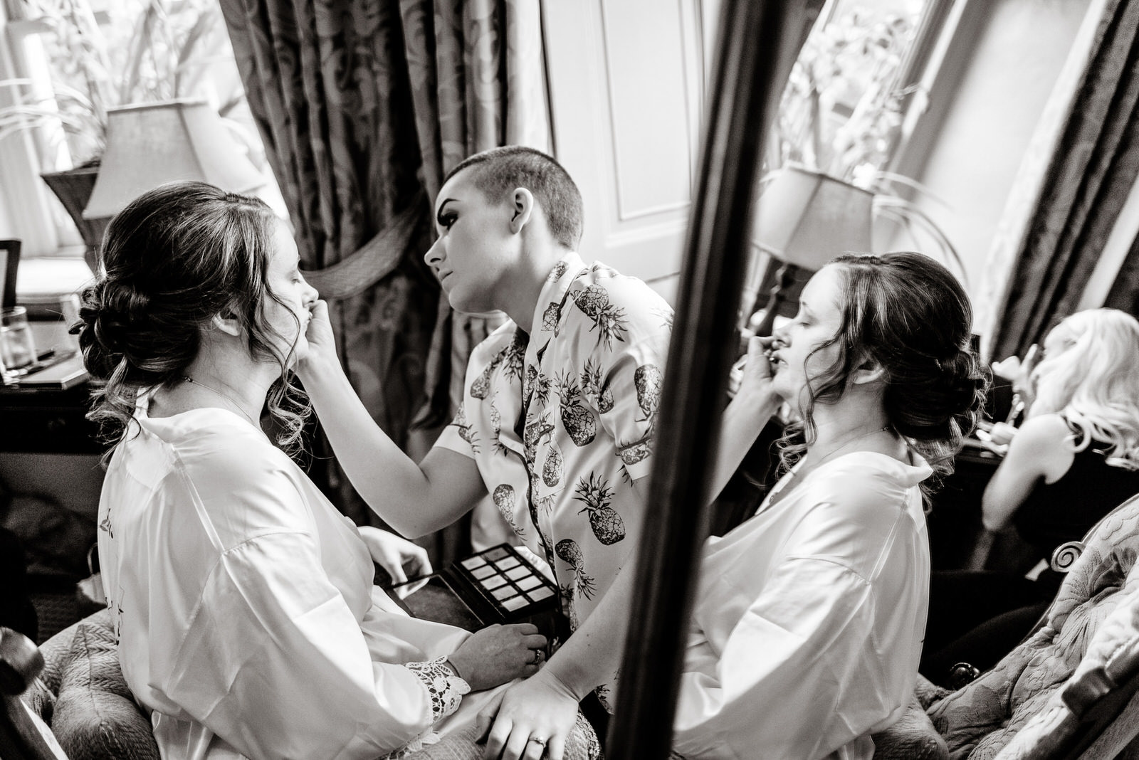 Two people in robes have makeup applied by an individual holding a palette, possibly preparing for a Washing Borough Hall wedding. They're seated near a window with curtain drapes, surrounded by makeup tools, while a child lingers in the background.