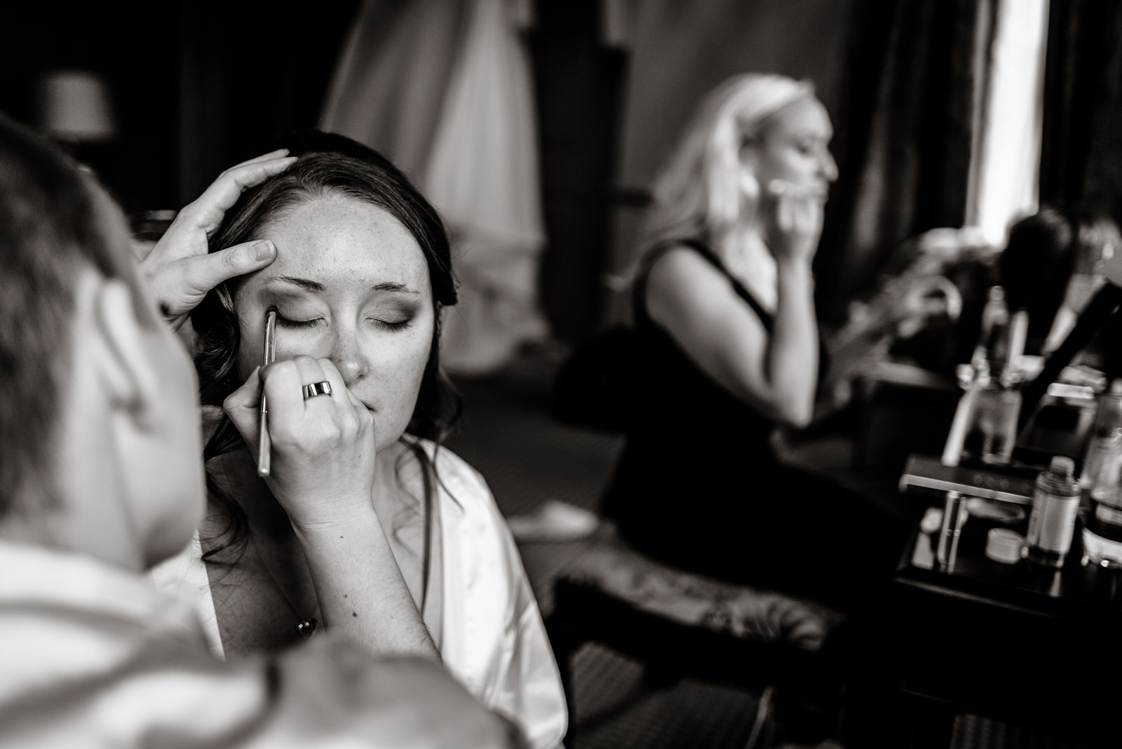 A woman has her eye makeup artfully applied in preparation for a wedding, embodying timeless beauty, while another woman in the background applies wa-inspired makeup in front of a mirror.