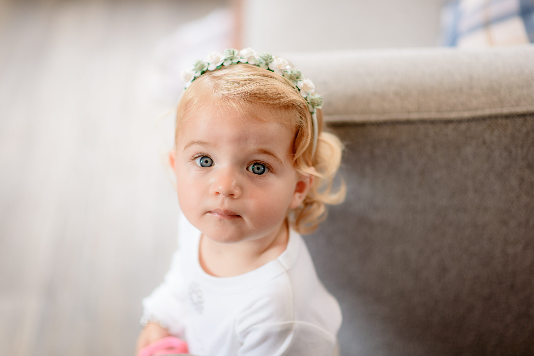 Toddler with blonde hair and a floral headband looks up, wearing a white shirt, as if envisioning a grand moment at Kenwick Park Hotel wedding, next to a gray couch.