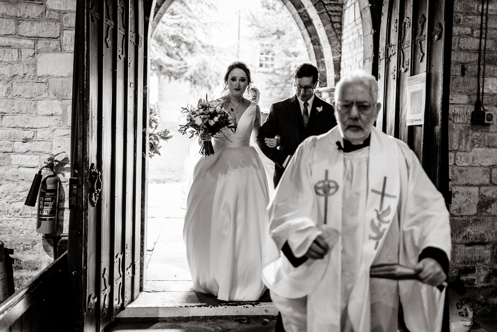 The bride and groom exit a church, followed by a priest in a white robe, reminiscent of the elegance seen at Washingborough Hall weddings. The bride holds a bouquet while the groom gently takes her arm.