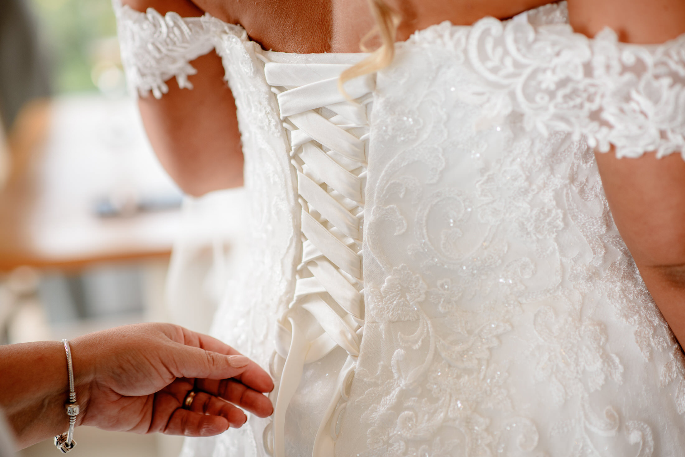 At a Kenwick Park Hotel wedding, a person carefully adjusts the laces on the back of an exquisite white lace wedding dress with off-the-shoulder sleeves.