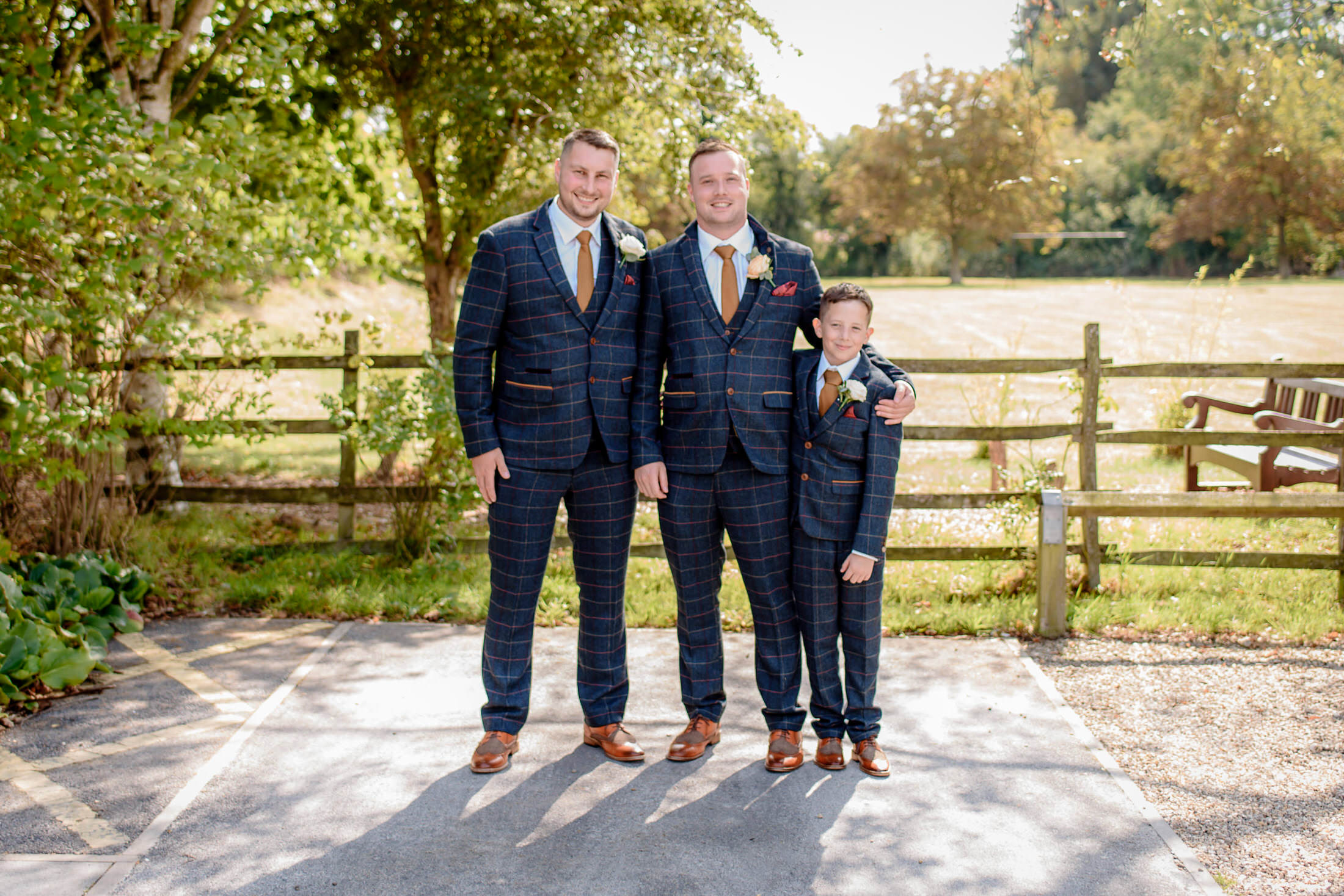 Three people in matching plaid suits stand outside the Brackenborough Hotel on a sunny day, with trees and a fence providing a picturesque backdrop.