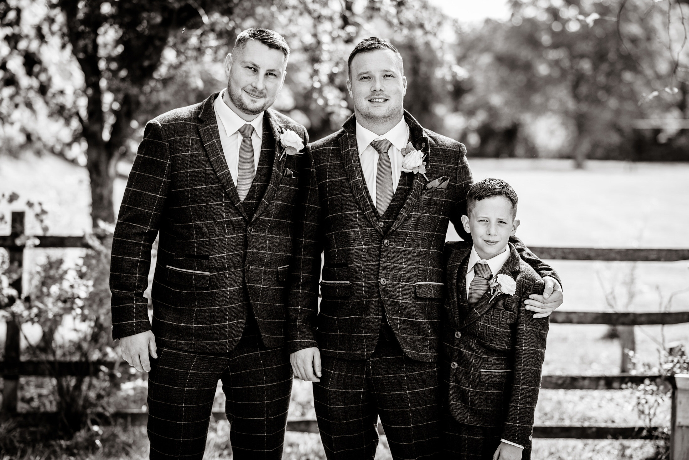 Three people in matching checkered suits and ties stand together outdoors, two adults and a child, with lush greenery in the background, evoking the elegance of a Kenwick Park Hotel wedding.