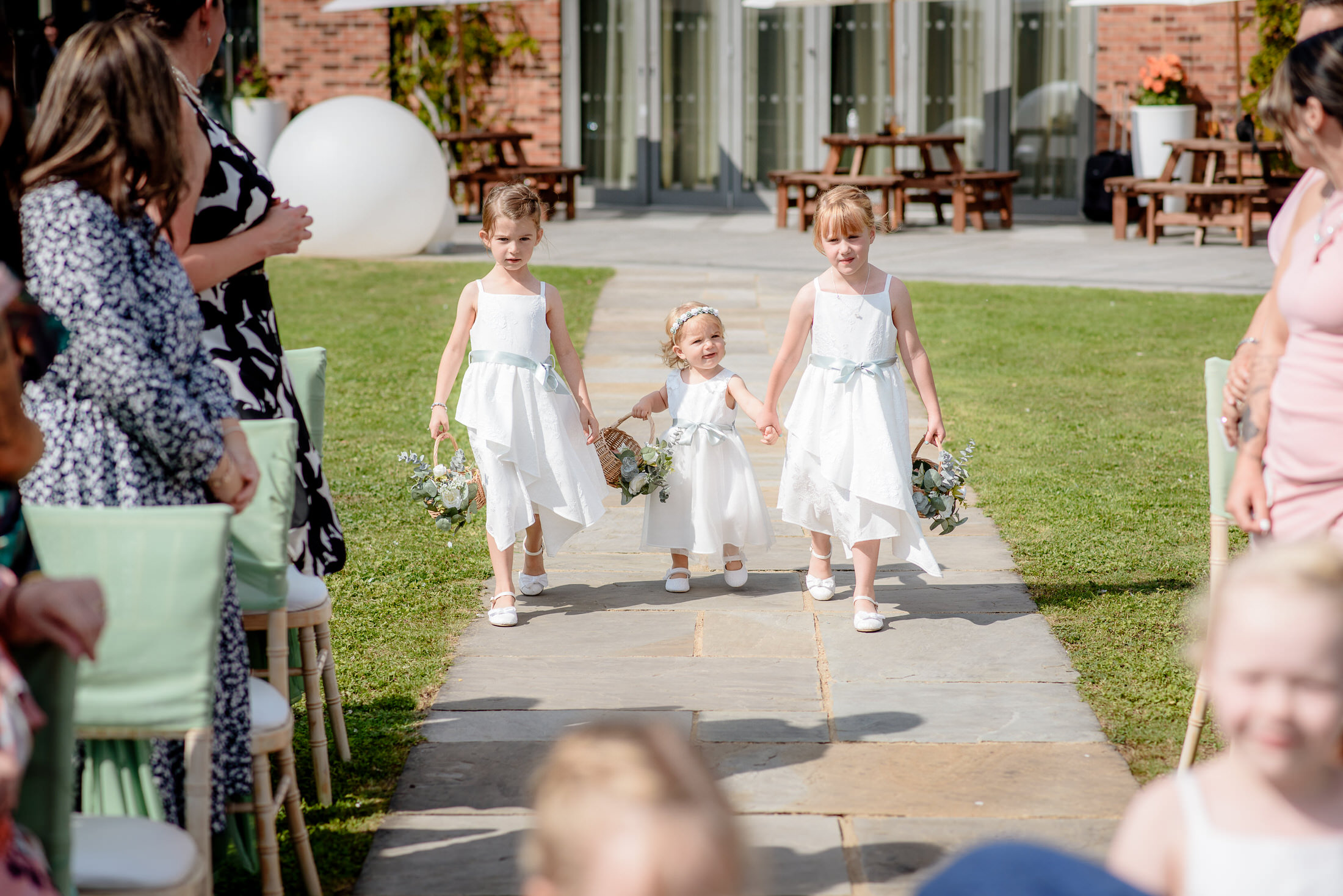 Three children in white dresses walk down an outdoor aisle holding flowers, surrounded by seated guests, at the charming Brackenborough Hotel.