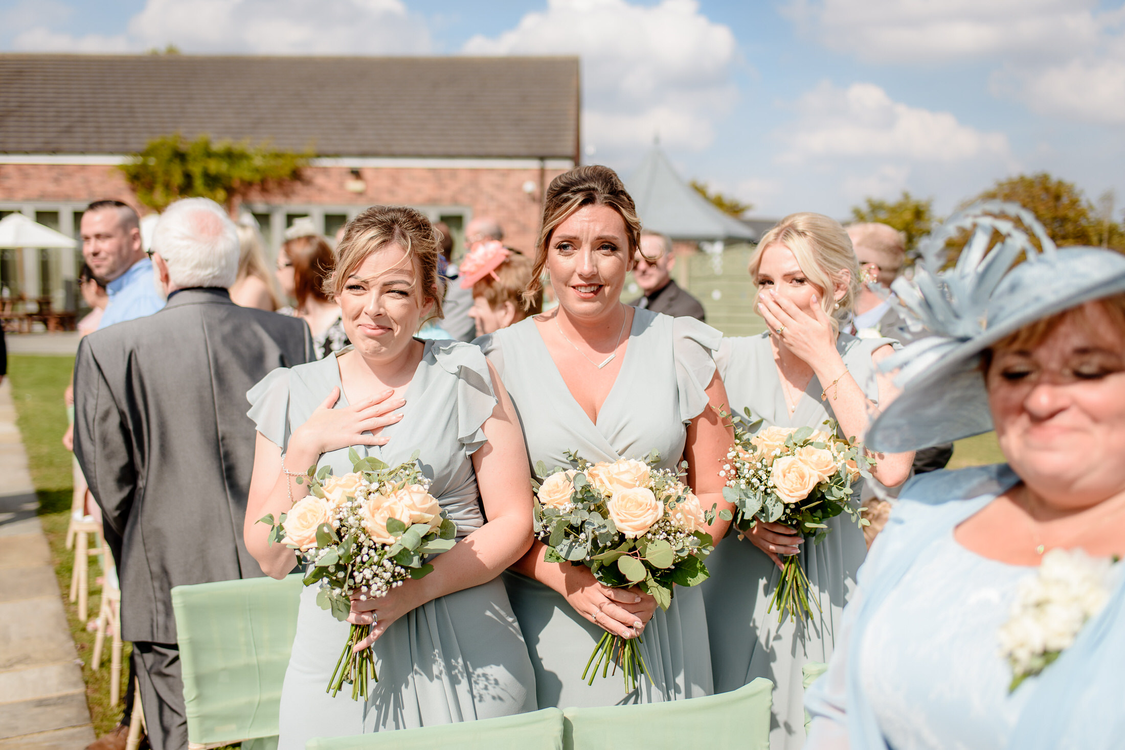 Three bridesmaids in light blue dresses holding bouquets stand outdoors at a wedding reception at Brackenborough Hotel. A woman in a hat stands in the foreground, slightly out of focus, with a charming building and picturesque sky completing the serene backdrop.