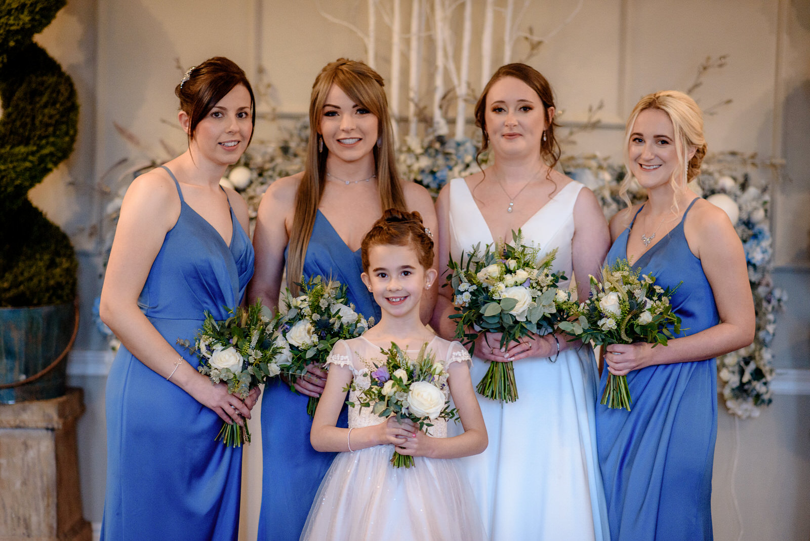 Five women in blue and white dresses holding bouquets, posing indoors at Washingborough Hotel. One is a child in a light pink dress. Floral arrangements and decorative plants are in the background, adding a touch of elegance to the wedding atmosphere.