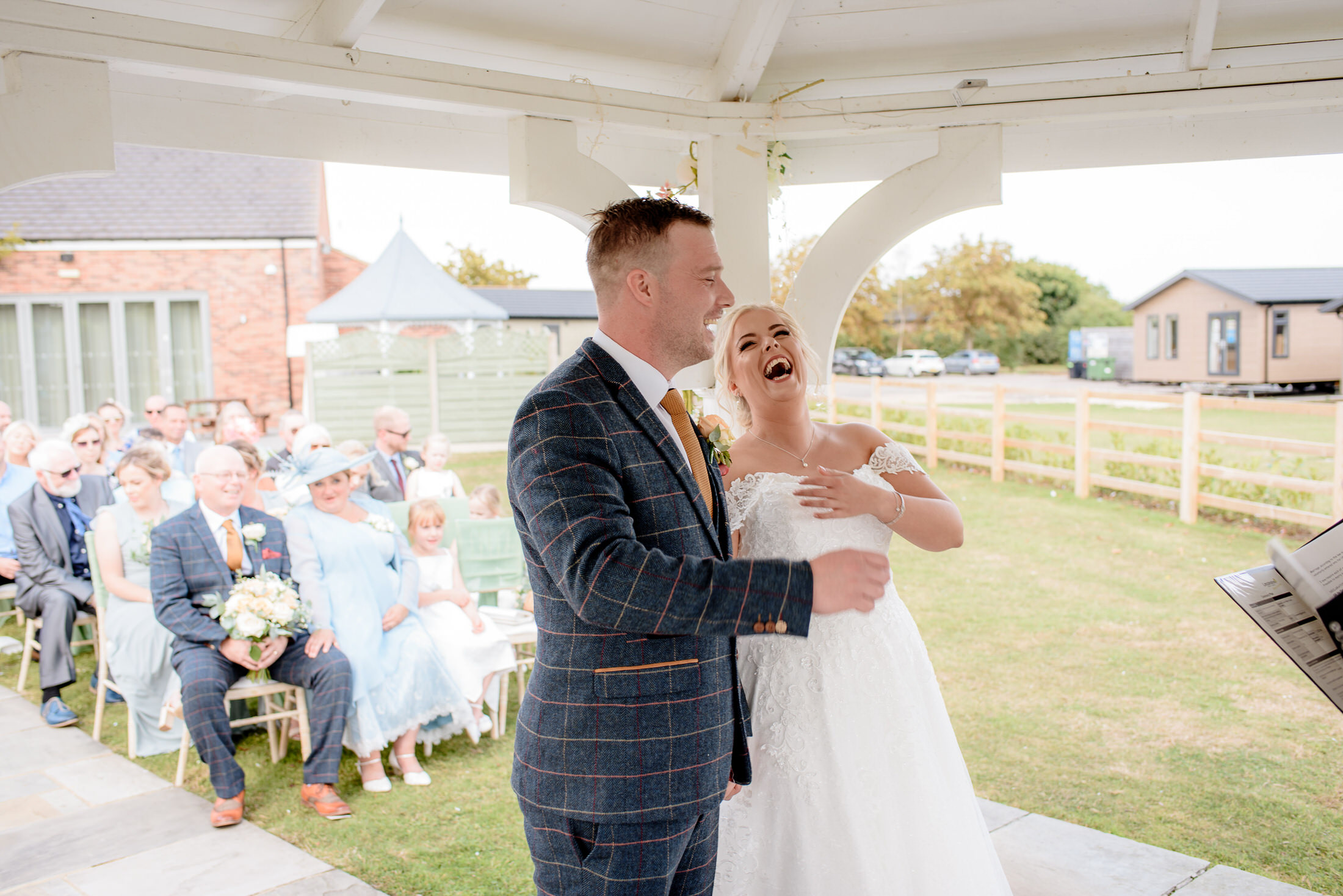 A couple stands at an outdoor wedding ceremony under a gazebo at Brackenborough Hotel. The bride laughs joyfully as the groom looks forward, with guests seated in the picturesque background.