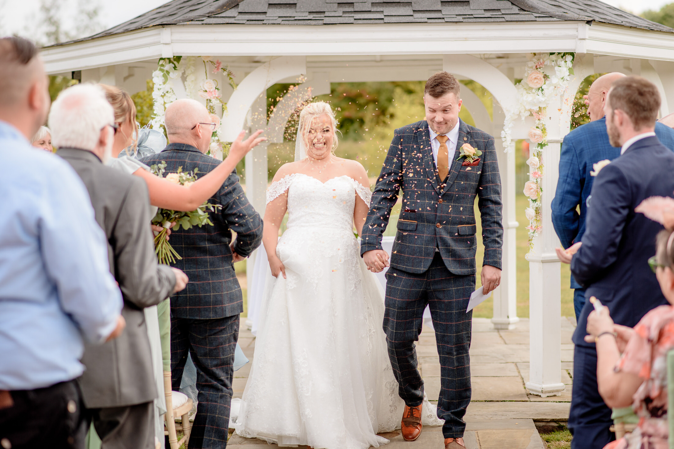 A newlywed couple walks down the aisle outdoors at Brackenborough Hotel, smiling as guests throw confetti. They are near a gazebo delicately decorated with flowers.