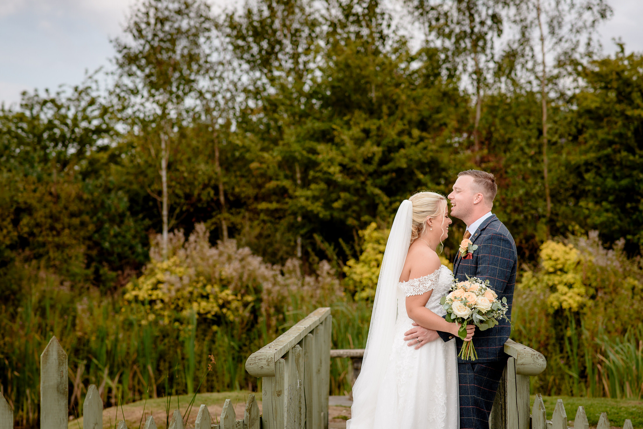 A couple embraces on a wooden bridge in the garden of Brackenborough Hotel. The bride, holding a bouquet, wears a white gown, while the groom is in a checked suit. Lush greenery surrounds them in this picturesque setting.