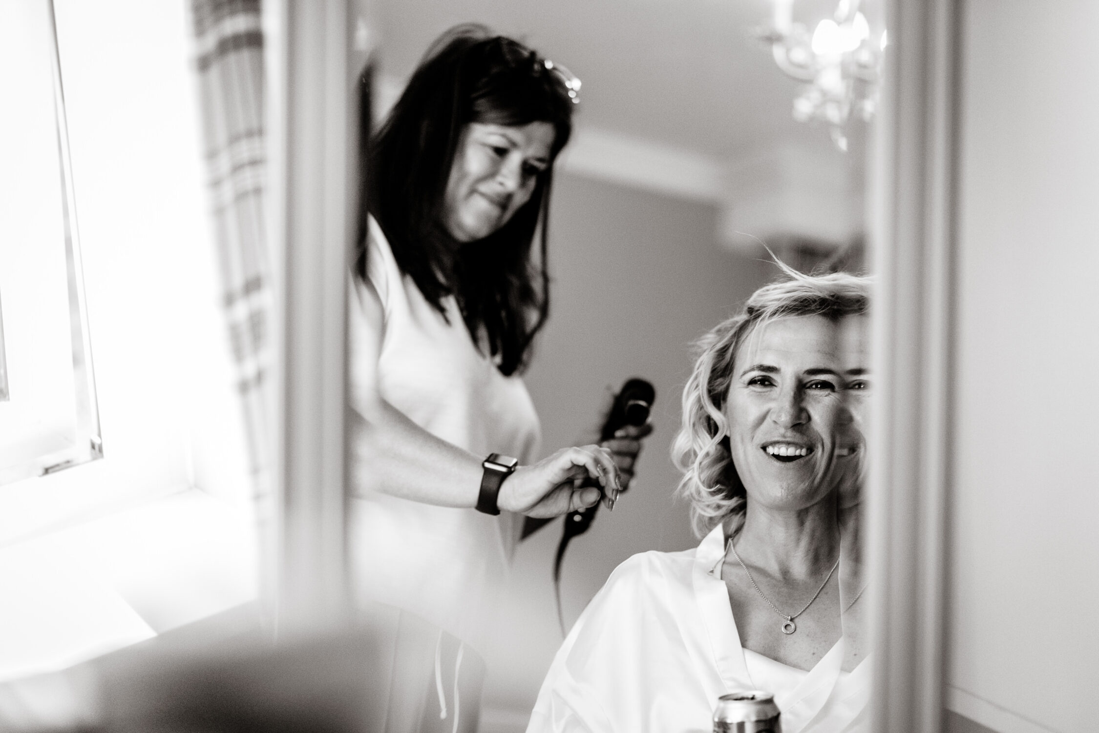 A woman smiles while having her hair styled by another woman, both visible in a mirror’s reflection, as they prepare for a Lincolnshire farm wedding.
