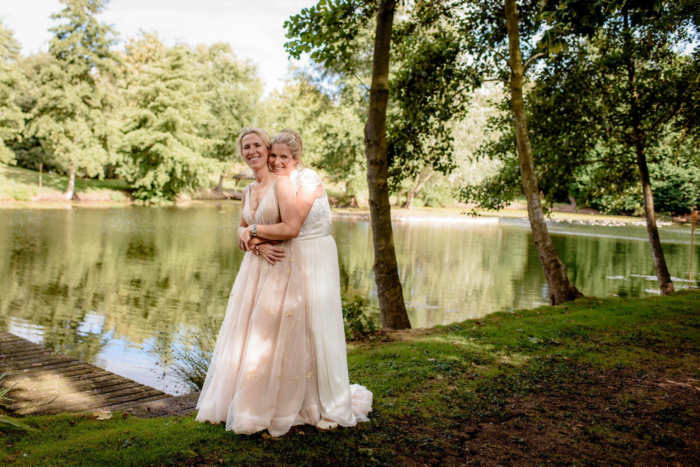 Two women in light-colored dresses embrace and smile while standing near a tree-lined lake on a sunny day, capturing the joy of a beautiful Lincolnshire farm wedding.