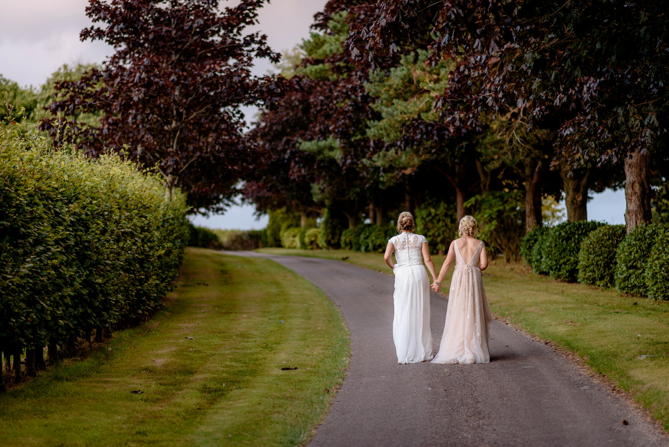 Two women in formal dresses walk hand in hand down a tree-lined path, surrounded by grass and greenery, with their backs to the camera—capturing a tender moment at a Lincolnshire Farm Wedding.