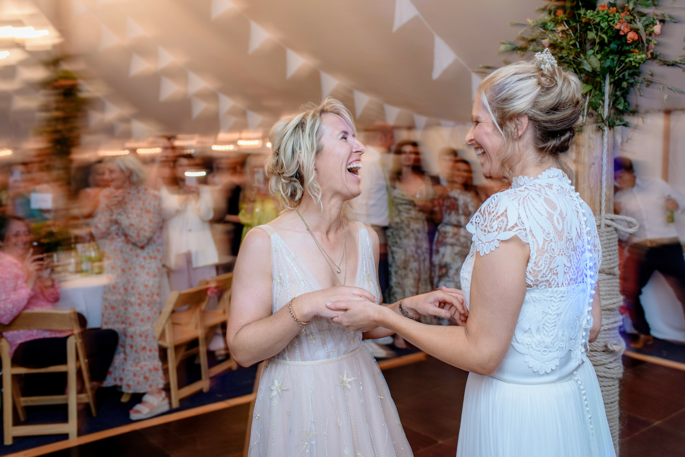 Two women in wedding dresses smile and hold hands while standing together at their Lincolnshire farm wedding reception, with guests blurred in the background.