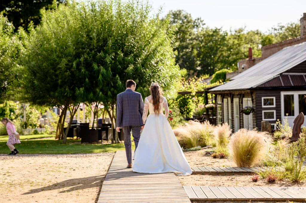 Lydia & Jonathan, a bride and groom, walk hand in hand along a wooden path in the Walled Garden Baumber, with a rustic building and trees creating the perfect wedding backdrop.
