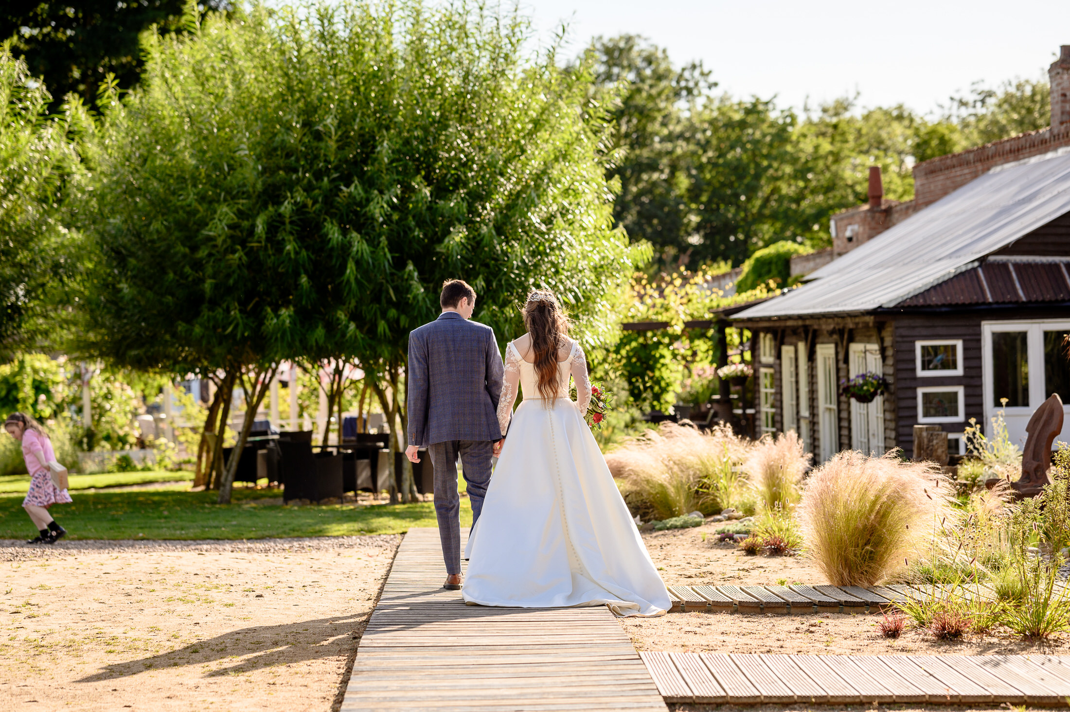 Lydia & Jonathan, a bride and groom, walk hand in hand along a wooden path in the Walled Garden Baumber, with a rustic building and trees creating the perfect wedding backdrop.