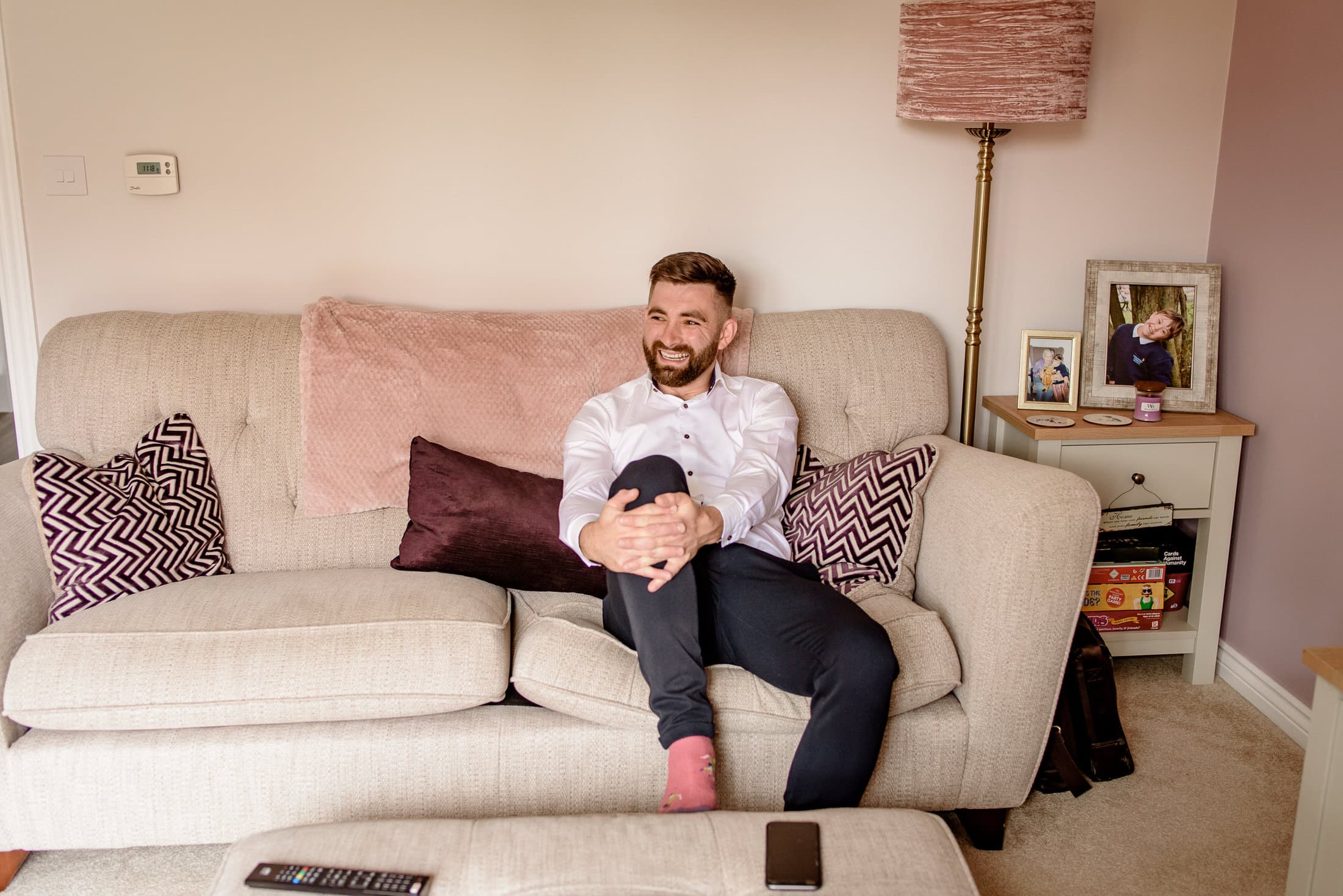 A man sits on a beige sofa, smiling with his hands round his knees. A remote and phone are on the coffee table, whilst family photos and a lamp evoke memories of a joyful Brackenborough Hotel wedding in the background.