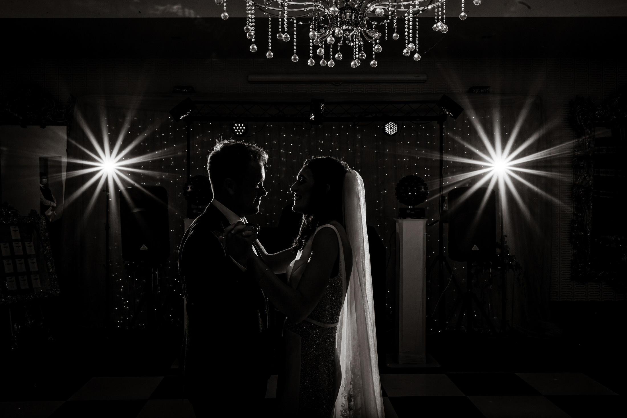 A bride and groom share a romantic dance beneath a chandelier, backlit by two bright lights in the dimly lit Brackenborough Hotel wedding venue.